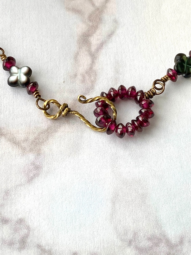 May include: A close-up of a necklace with a brass clasp and a circle of dark red beads. The necklace also features a small white flower bead.
