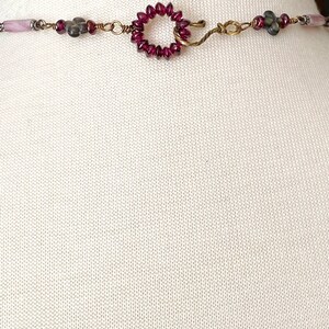 May include: A close-up of a necklace clasp with a circle of small red beads and a hook closure. The necklace is made with wire and has other beads in shades of gray and pink.