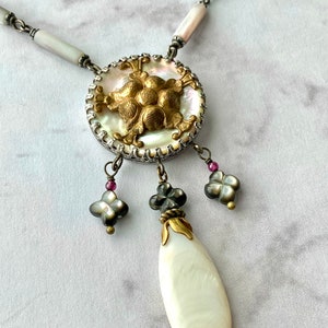 May include: A white and gold necklace with a large, round pendant featuring a floral design. The pendant is accented with small, black and white beads and a teardrop-shaped pearl.