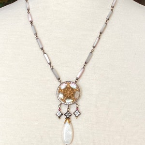 May include: A white and gray shell necklace with a round pendant featuring a gold and white shell design. The pendant has a pearlescent drop hanging from it.