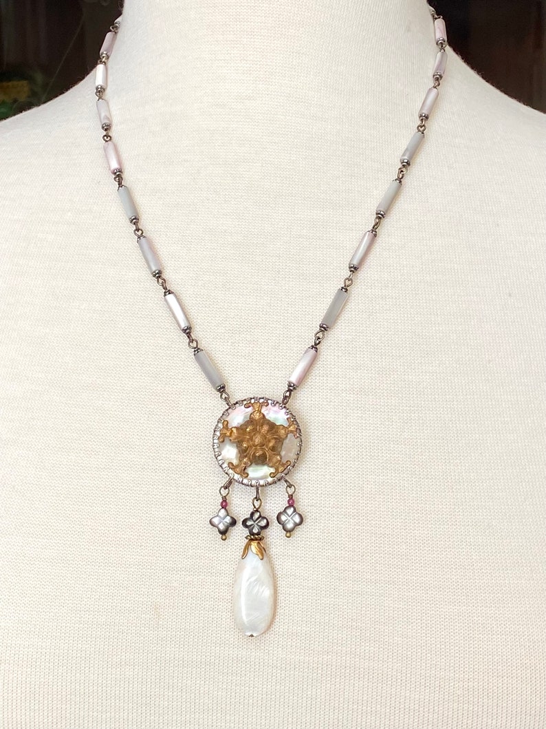 May include: A white and gray shell necklace with a round pendant featuring a gold and white shell design. The pendant has a pearlescent drop hanging from it.