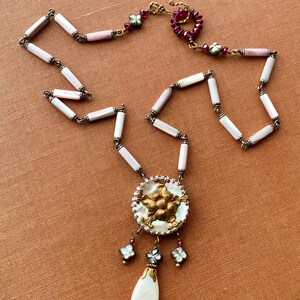 May include: A white and pink shell necklace with a gold and white pendant featuring a floral design. The pendant is attached to a teardrop-shaped white shell. The necklace has a red and gold clasp.
