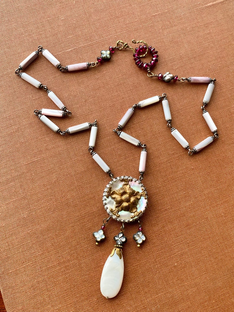 May include: A white and pink shell necklace with a gold and white pendant featuring a floral design. The pendant is attached to a teardrop-shaped white shell. The necklace has a red and gold clasp.