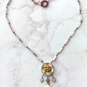 May include: A delicate necklace with a white and pink shell bead chain and a gold and white pendant with a floral design. The pendant has a white teardrop-shaped bead hanging from it.