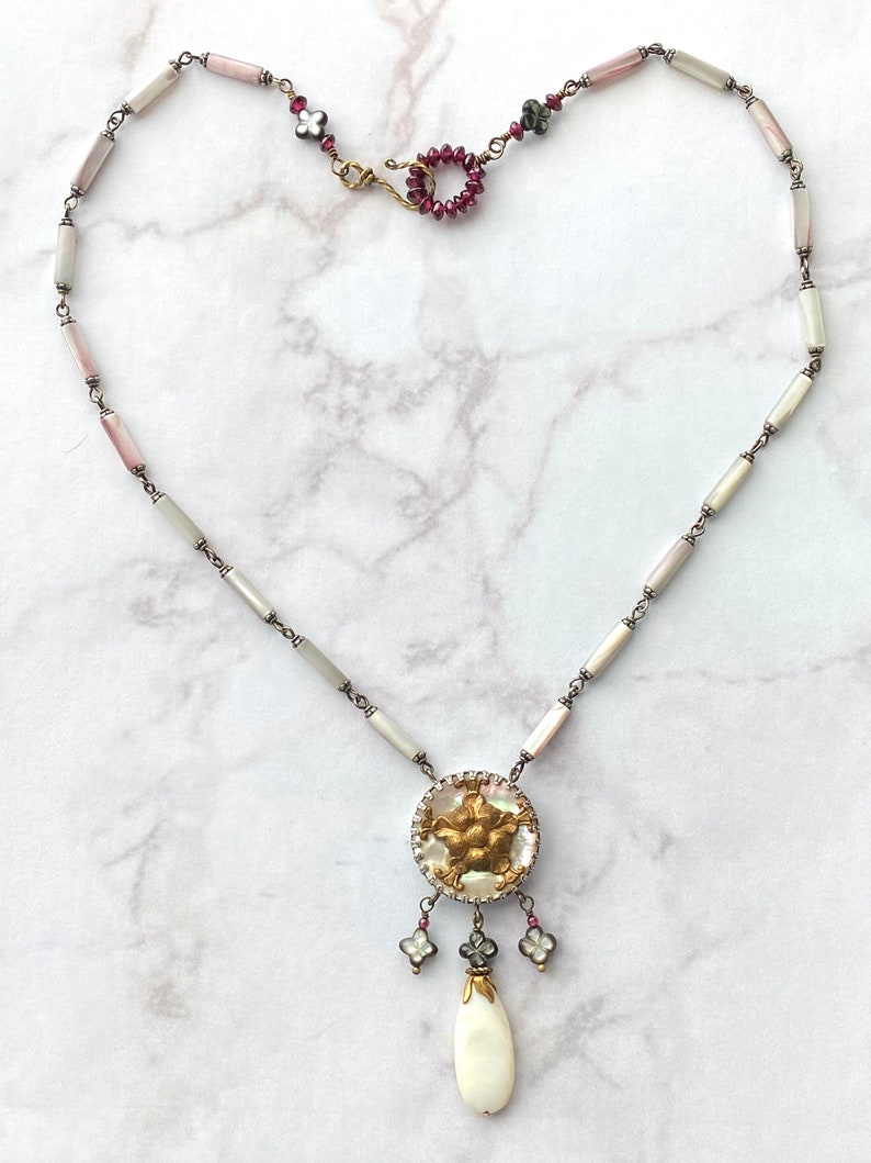 May include: A delicate necklace with a white and pink shell bead chain and a gold and white pendant with a floral design. The pendant has a white teardrop-shaped bead hanging from it.