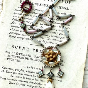 May include: A white and gold necklace with a large, round pendant featuring a floral design. The pendant is made of mother-of-pearl and has a gold-tone finish. The necklace is made of white and pink beads and has a silver-tone chain. The necklace is hanging on a page of a book with text that reads "SIXEME INTE MEDE. F F DES JEUX THIENS."