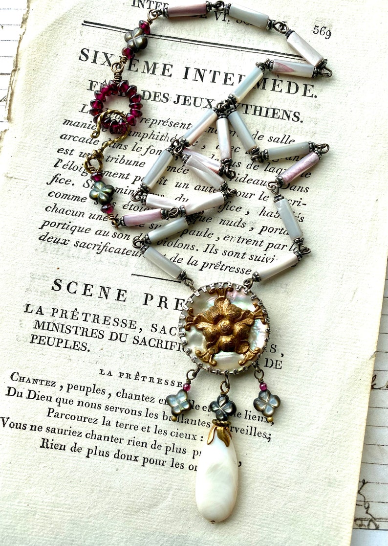 May include: A white and gold necklace with a large, round pendant featuring a floral design. The pendant is made of mother-of-pearl and has a gold-tone finish. The necklace is made of white and pink beads and has a silver-tone chain. The necklace is hanging on a page of a book with text that reads "SIXEME INTE MEDE. F F DES JEUX THIENS."