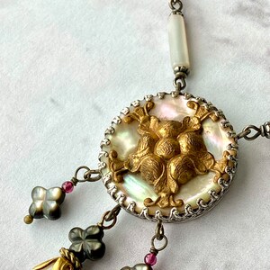 May include: A silver and gold necklace with a large, round, iridescent mother-of-pearl pendant. The pendant features a gold, ornate design and is surrounded by a silver chain. The necklace also has three smaller, black, clover-shaped pendants with gold accents.