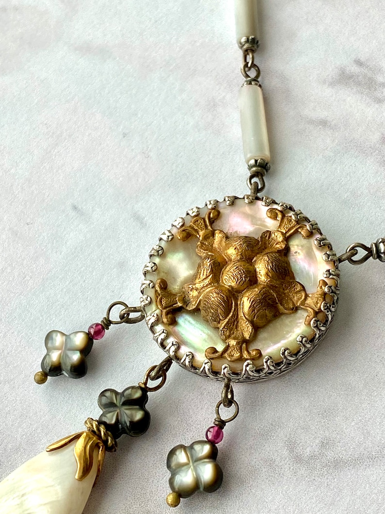 May include: A silver and gold necklace with a large, round, iridescent mother-of-pearl pendant. The pendant features a gold, ornate design and is surrounded by a silver chain. The necklace also has three smaller, black, clover-shaped pendants with gold accents.