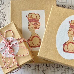 May include: Three brown gift boxes with white labels. The labels feature a vintage illustration of a woman wearing a crown. The woman is depicted in profile and has a red and pink color scheme.