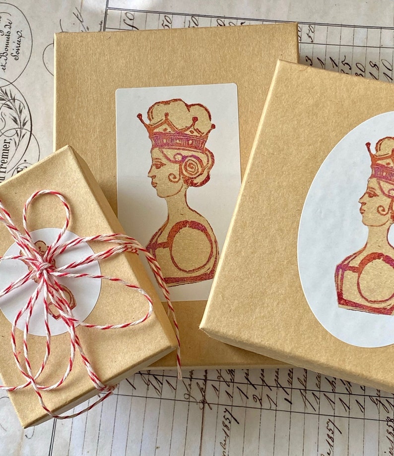 May include: Three brown gift boxes with white labels. The labels feature a vintage illustration of a woman wearing a crown. The woman is depicted in profile and has a red and pink color scheme.