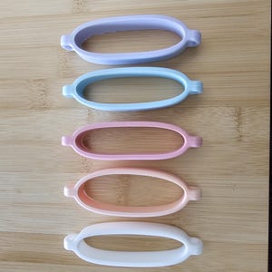 May include: Five oval silicone bands in pastel shades: lilac, pale blue, pink, peach, and white. Each band features small, rounded tabs on either side. The bands are arranged vertically on a wooden surface.
