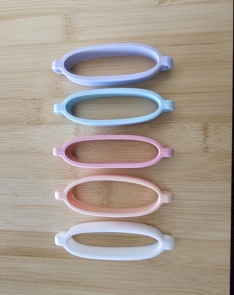 May include: Five oval silicone bands in pastel shades: lilac, pale blue, pink, peach, and white. Each band features small, rounded tabs on either side. The bands are arranged vertically on a wooden surface.
