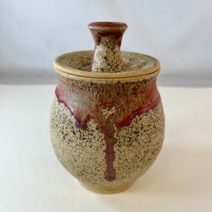 May include: A small, speckled ceramic jar with a lid. The jar is a light beige color with brown speckles and a red and brown glaze that drips down the side. The lid is a light beige color with a brown rim.