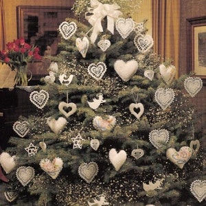 May include: A decorated Christmas tree with heart-shaped ornaments. The tree is adorned with white lace and fabric hearts, stars, and angel ornaments. A large white bow sits at the top, and the tree is surrounded by greenery and small white flowers.