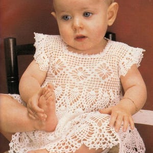 Puede incluir: Un bebé con un vestido blanco de ganchillo hecho a mano con un diseño floral. El vestido tiene mangas cortas y un dobladillo festoneado. El bebé, con ojos azules, está sentado en una silla.
