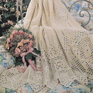 May include: A crocheted throw blanket in a cream color with a delicate openwork pattern. The blanket is draped over a bed with floral patterned bedding and a bouquet of pink flowers.