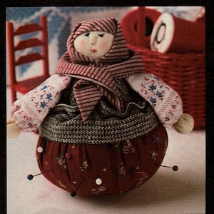 May include: A red and white striped fabric pincushion shaped like a woman wearing a red and white striped scarf and a white blouse with floral embroidery. The pincushion is filled with pins and is sitting on a white surface.