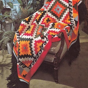 May include: A colorful, geometric patterned blanket draped over a vintage armchair. The blanket features a vibrant design with red, orange, black, white, and green hues. It has a fringed edge and is displayed in a well-lit room with plants.