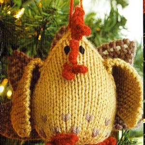 May include: A knitted chicken Christmas tree decoration. The ornament is yellow with red detailing for the comb, beak, and feet. It has brown wings and black eyes. It hangs from a red string, and is surrounded by green foliage.
