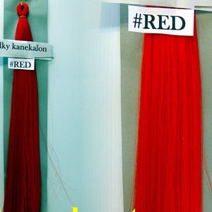 May include: Two samples of bright red silky kanekalon hair extensions. The text "Silky kanekalon" and "#RED" are printed on white labels attached to the hair. The hair is hanging against a light blue background.