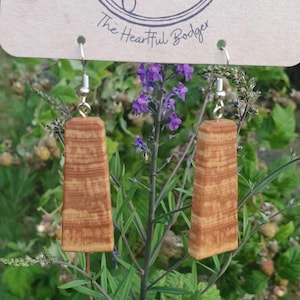 May include: Wooden rectangular earrings with a natural wood grain pattern, suspended from silver-coloured hooks. The background features green foliage and purple flowers. A tag above the earrings reads "The Heartful Bodger".