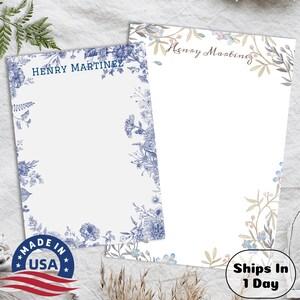 May include: Two personalized stationery notepads with floral borders. The notepad on the left has a blue floral border and the name "HENRY MARTINEZ" printed at the top. The notepad on the right has a light blue and brown floral border and the name "Henry Martinez" printed at the top. Both notepads have a white background.
