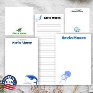 May include: Four different notepads with lined paper and a variety of sea-themed illustrations. The notepads feature the name "Kevin Moore" and the text "A Note From". One notepad has a green turtle, one has a blue whale, one has a blue jellyfish, and one has a blue seashell.