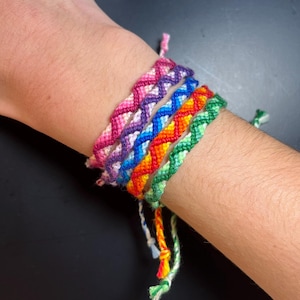 May include: A set of six friendship bracelets in a rainbow color pattern. The bracelets are made with a chevron stitch and have white string ties.