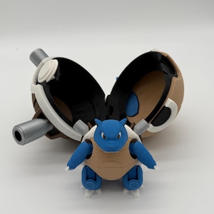 May include: A blue and tan Blastoise figurine standing in front of an open, spherical, tan and white container with two silver-colored cylindrical protrusions on the sides. The interior of the container is black with a blue circular accent.