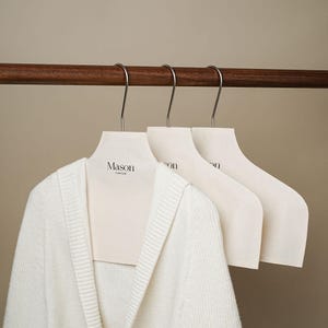 May include: Three off-white clothing hangers with metal hooks hang from a wooden rod. The first hanger holds a white cardigan. The hangers have the word "Mason" printed on them in black, with the words "Cute Crush" below.