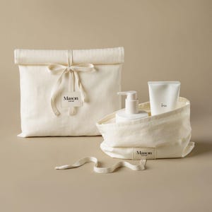 May include: Two off-white canvas bags with roll-top closures and tie ribbons. One bag is closed with a Mason label, while the other is open, containing a white pump bottle and a white squeeze tube labelled IPSA, with a Mason label on the front.