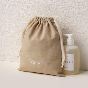 Puede incluir: Una bolsa beige con cordón con el texto "Mason Cute Craft" y una botella blanca con dispensador de bomba. La bolsa está hecha de una tela texturizada. La botella tiene una etiqueta blanca con texto. Los artículos son probablemente parte de un set de regalo o un producto para el cuidado de la piel.
