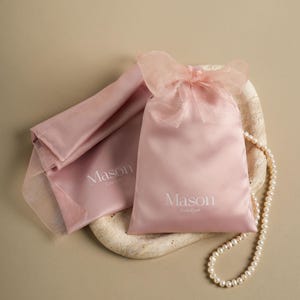 Puede incluir: Dos bolsas de sat&eacute;n rosa p&aacute;lido, una con un lazo transparente, con la palabra "Mason" impresa. Un collar de perlas descansa sobre una superficie con aspecto de piedra. Las bolsas son para joyas o peque&ntilde;os regalos.
