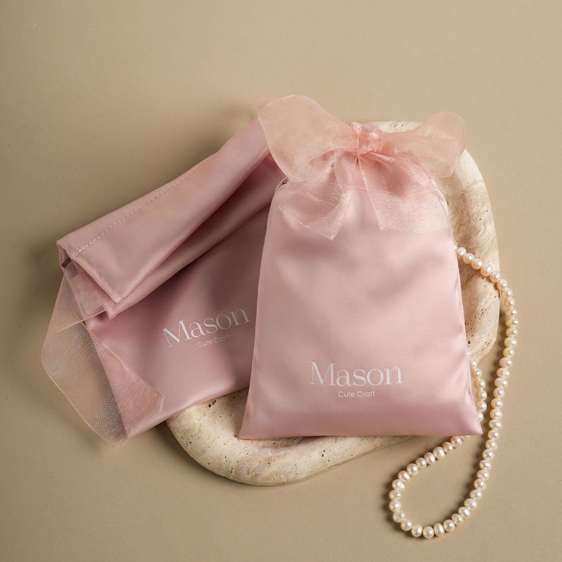 Puede incluir: Dos bolsas de sat&eacute;n rosa p&aacute;lido, una con un lazo transparente, con la palabra "Mason" impresa. Un collar de perlas descansa sobre una superficie con aspecto de piedra. Las bolsas son para joyas o peque&ntilde;os regalos.