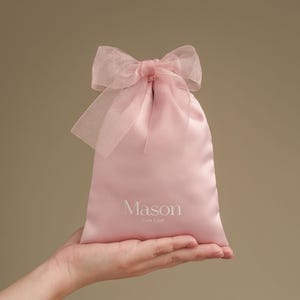 Puede incluir: Una bolsa de regalo de sat&eacute;n rosa claro con un lazo transparente en la parte superior. La bolsa tiene el nombre "Mason" y las palabras "Cute Craft" impresas en blanco. La bolsa est&aacute; sostenida en una mano.