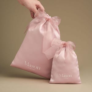 Puede incluir: Dos bolsas rosas de sat&eacute;n con lazos de cinta transparente. La bolsa m&aacute;s grande tiene la palabra "Mason" y "Cute Craft" impresa en blanco. La bolsa m&aacute;s peque&ntilde;a tambi&eacute;n tiene la palabra "Mason" y "Cute Craft" impresa en blanco.