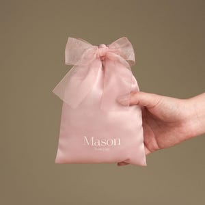 Puede incluir: Una bolsa de regalo de sat&eacute;n rosa claro con un lazo transparente. La bolsa est&aacute; sujeta por una mano y presenta el nombre "Mason" y las palabras "Cute Craft" impresas en blanco.