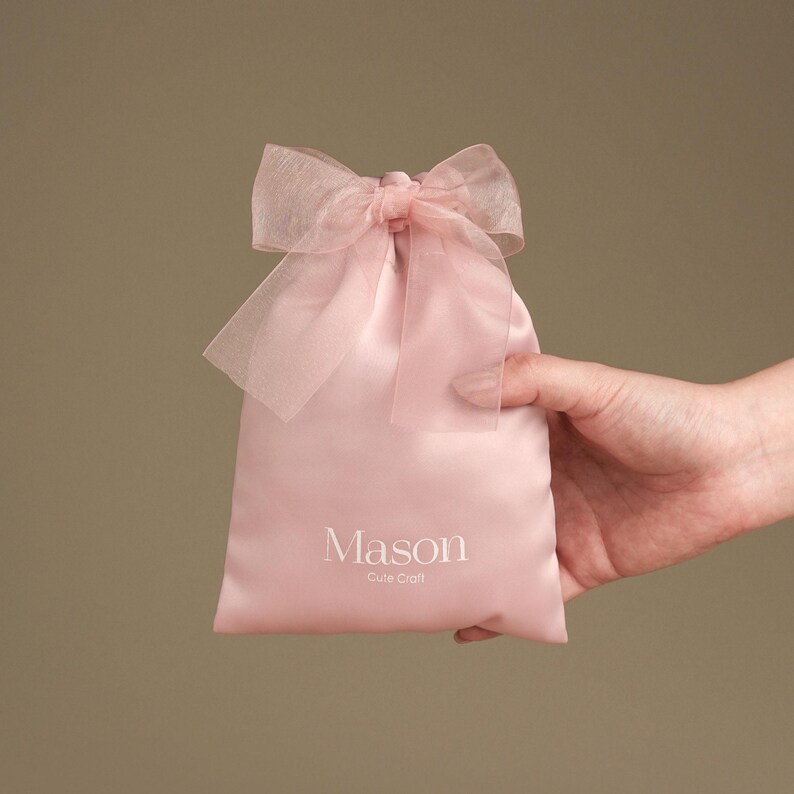 Puede incluir: Una bolsa de regalo de sat&eacute;n rosa claro con un lazo transparente. La bolsa est&aacute; sujeta por una mano y presenta el nombre "Mason" y las palabras "Cute Craft" impresas en blanco.