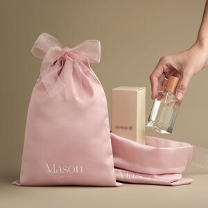 Puede incluir: Una bolsa de sat&eacute;n rosa claro con un lazo transparente, una caja a juego y un frasco de perfume con tap&oacute;n de madera. La bolsa tiene el texto "Mason Cute Craft" impreso.