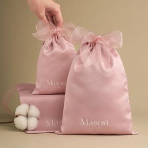 Puede incluir: Bolsas rosas de sat&eacute;n con lazos de cinta transparente. Las bolsas tienen la palabra "Mason" impresa en blanco. Una ramita de algod&oacute;n est&aacute; en primer plano. Probablemente para regalos o embalaje.