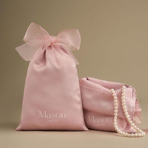Puede incluir: Dos bolsas de sat&eacute;n rosa p&aacute;lido con cord&oacute;n, una vertical con un lazo transparente, la otra abierta con un collar de perlas. Las bolsas tienen la palabra "Mason" y "Cute Craft" impresas en blanco.