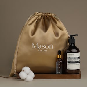以下が含まれることがあります： 「Mason Cute Craft」の文字が入ったゴールドサテン巾着袋が、黒いポンプ付きの茶色のボトル、小さなドロッパーボトル、綿球の隣にあります。ボトルは木製のトレイにあります。