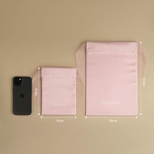 Puede incluir: Tres bolsas rosas de sat&eacute;n con cord&oacute;n en diferentes tama&ntilde;os, con la palabra "Mason" impresa. La bolsa m&aacute;s grande mide 30 cm de alto y 20 cm de ancho. La bolsa m&aacute;s peque&ntilde;a mide 18 cm de alto y 13 cm de ancho. Un smartphone negro est&aacute; al lado de la bolsa m&aacute;s peque&ntilde;a.