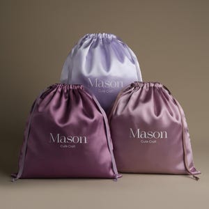 Könnte beinhalten: Drei Satin-Kordelzugbeutel in Lila-, Lavendel- und Mauve-Tönen. Jeder Beutel trägt den Namen "Mason" in einer weißen, eleganten Schrift, darunter "Cute Craft". Die Beutel sind oben mit passenden Satinbändern zusammengezogen.