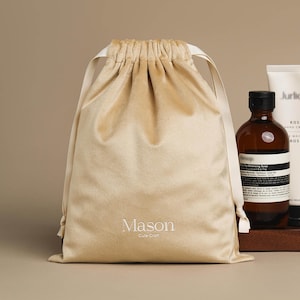 Pode incluir: Uma bolsa de veludo bege com cordão, com as palavras "Mason" e "Cute Craft" impressas em branco. A bolsa está ao lado de uma garrafa marrom com tampa preta e um tubo de loção branca.