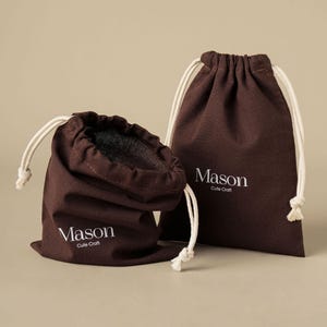 Può includere: Due sacchetti in tessuto marrone scuro con coulisse in corda bianca. Ogni sacchetto presenta la scritta bianca stampata Mason Cute Craft sul davanti. Un sacchetto è in posizione verticale mentre l'altro è ripiegato, rivelando la fodera interna.