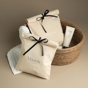 May include: Two white canvas bags with black ribbon ties and the text "Mason Cute Craft" printed on them. The bags are in a woven basket with a white tube of lotion.