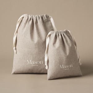 Könnte beinhalten: Zwei beigefarbene Kordelzugbeutel aus strukturiertem Stoff. Jeder Beutel hat weiße Baumwollkordeln und den in Weiß aufgedruckten Schriftzug Mason Cute Craft auf der Vorderseite. Die Beutel haben unterschiedliche Größen, der größere steht links.