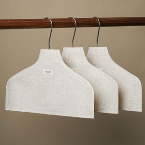 May include: Three off-white linen covered hangers with silver metal hooks hang from a dark brown wooden rod. The hangers have a wide, curved shape and a small label that reads "Mason". The background is a neutral beige.